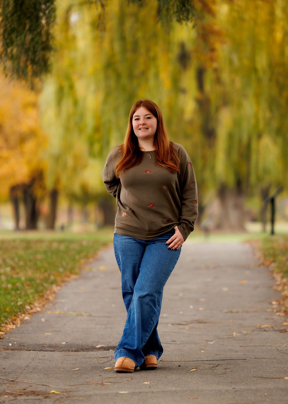 Someone walks along a peaceful autumn path with fallen leaves and willow trees creating a warm golden backdrop.
