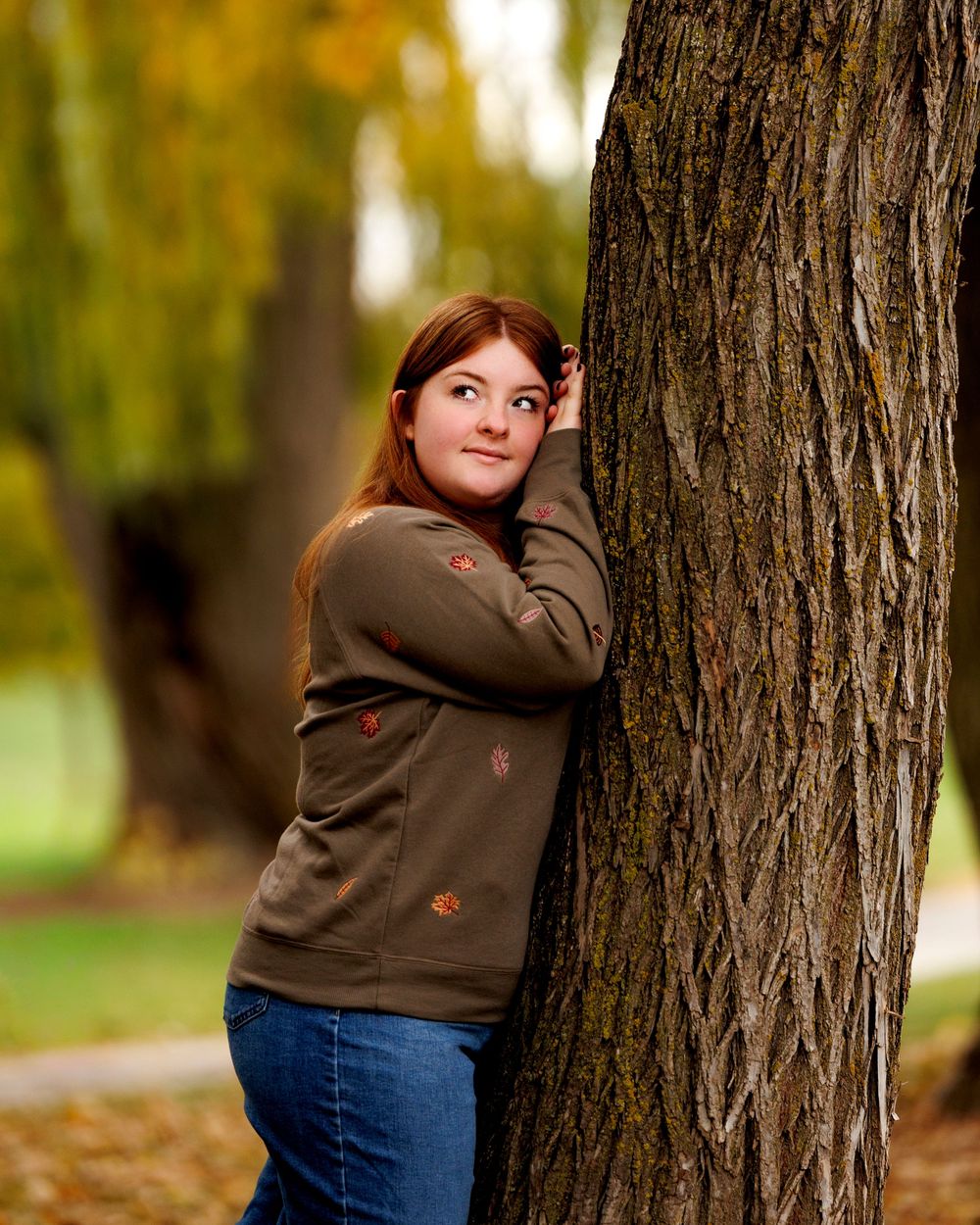 A person in a brown hoodie and jeans leans playfully against a large tree trunk in an autumn setting.