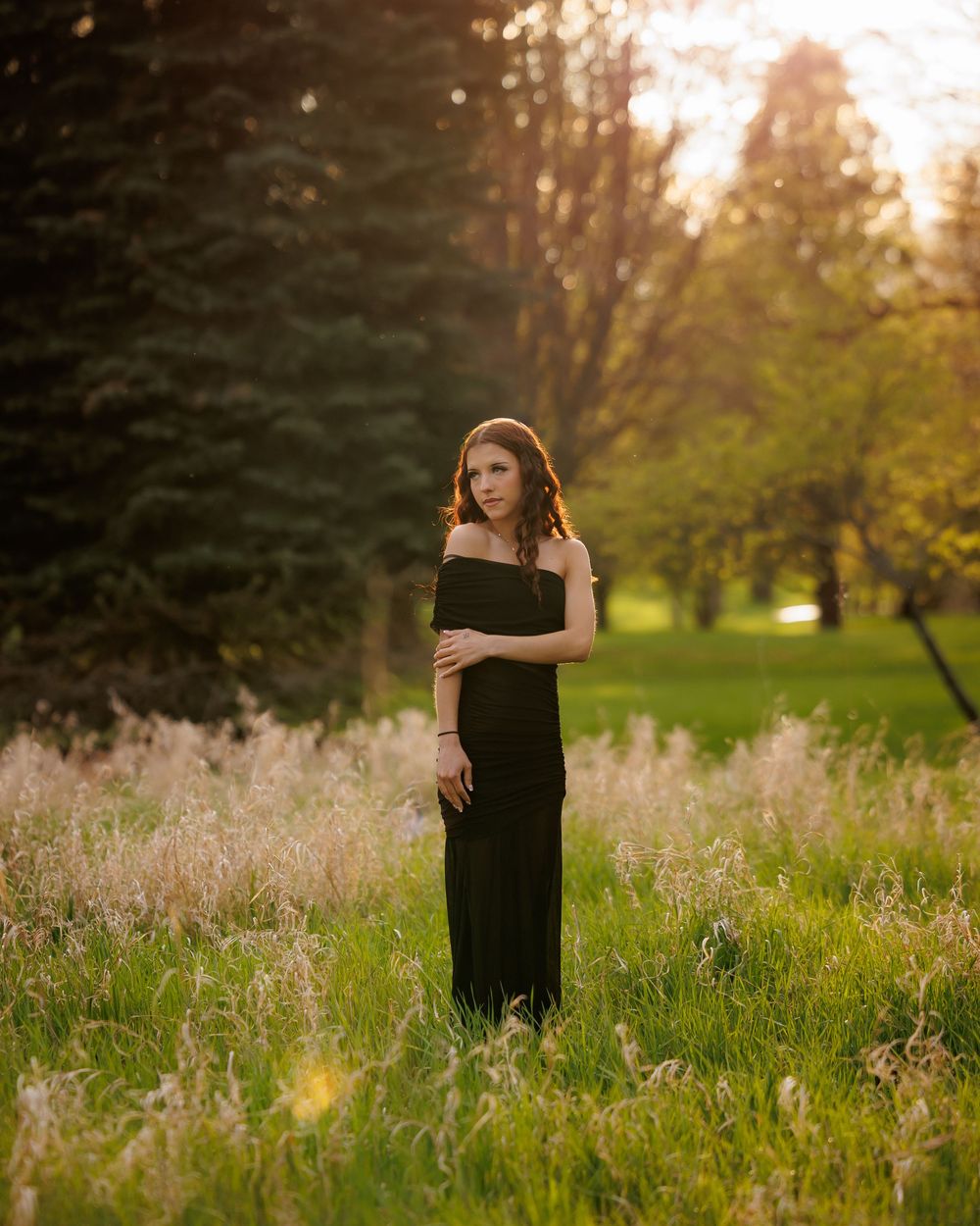 A woman in a black off-shoulder gown stands in a sunlit meadow surrounded by tall grass and trees at golden hour.