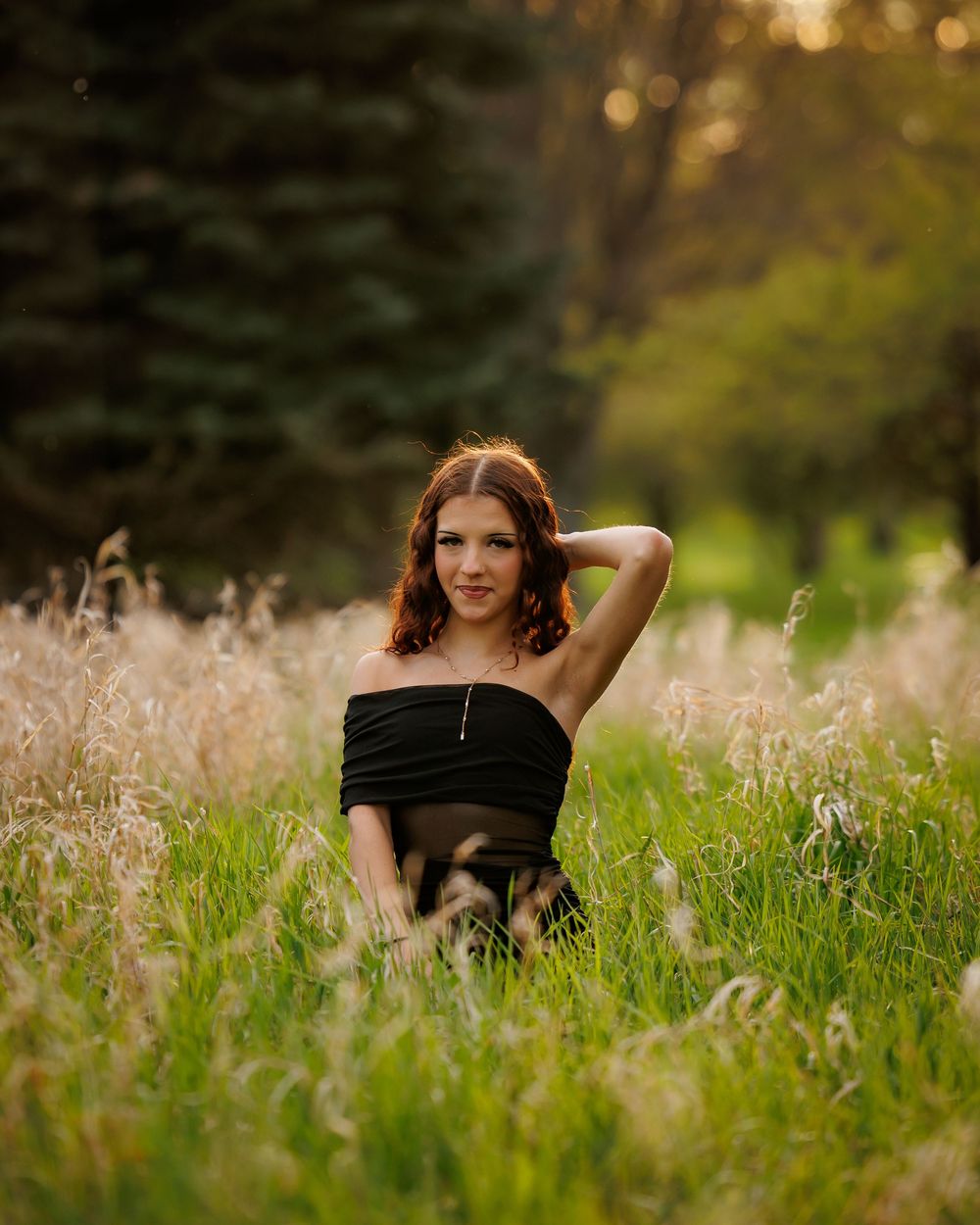 Young woman in black strapless dress sitting in tall grass field with trees in golden hour light.