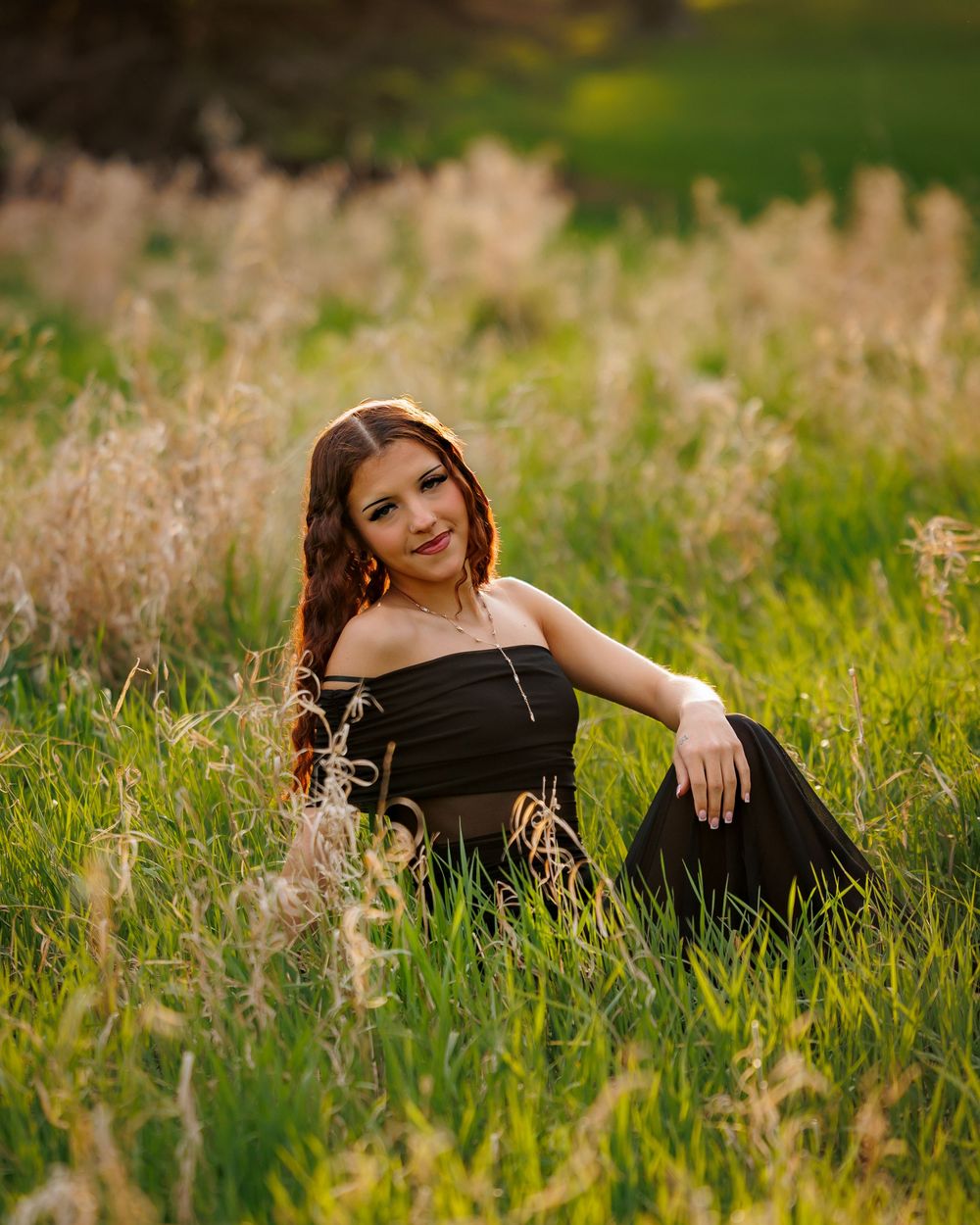 Young woman in black dress sitting gracefully in a lush green field with tall golden grasses during golden hour.