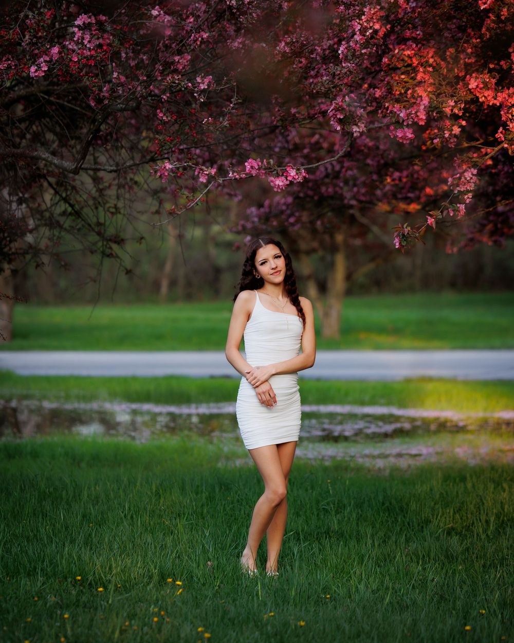 Young woman in white dress standing on green grass under blooming pink flowering tree in a park at dusk.