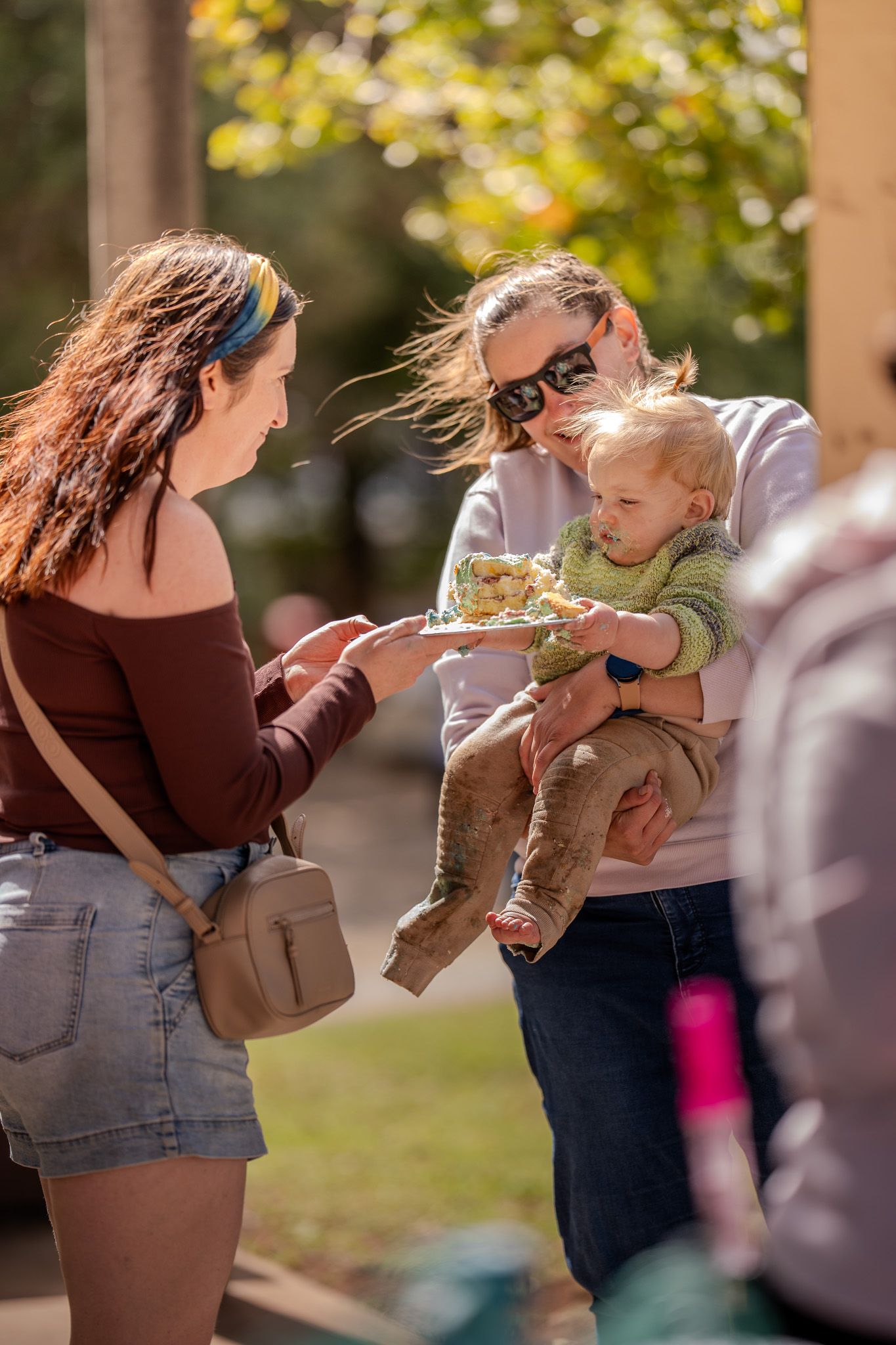 People enjoying a sunny autumn day outdoors while a small toddler in khaki pants is being held and fed.