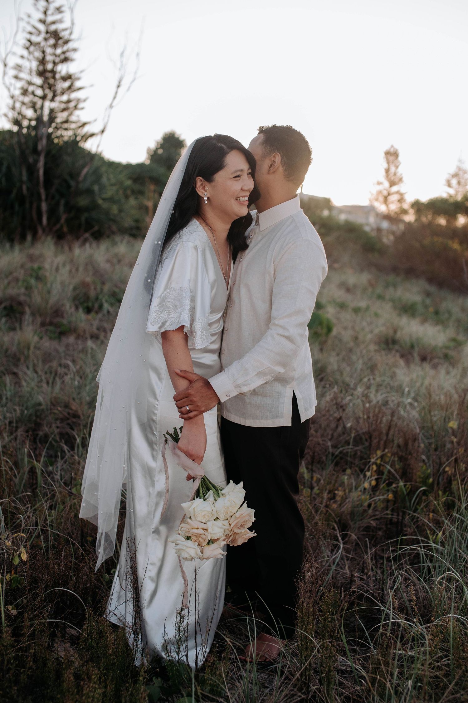 A newlywed couple sharing an intimate moment in a field at sunset while bride holds a white floral bouquet.