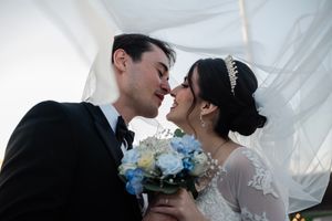 A romantic wedding moment under white fabric with a blue and white bouquet between the couple.