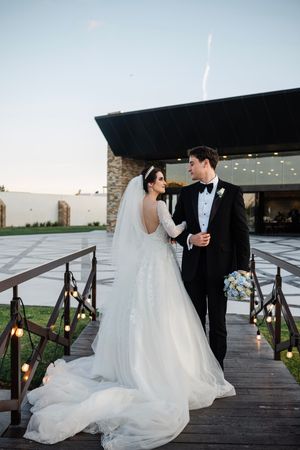 A romantic wedding couple shares an intimate moment on a rustic wooden deck with string lights overhead at dusk.