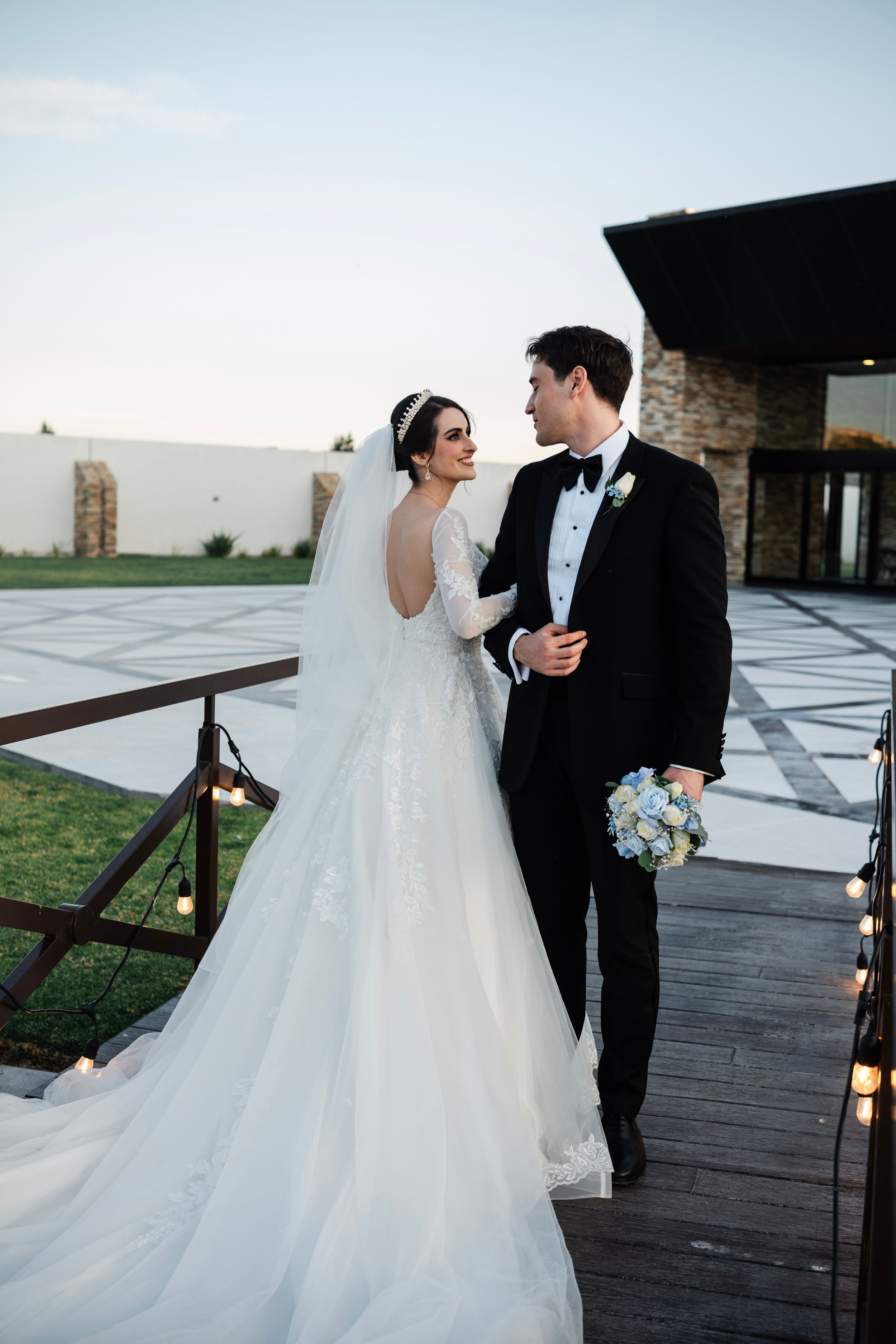 A bride in a flowing white gown and groom in black tuxedo share tender moments on an outdoor wooden deck.