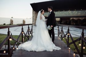 Romantic wedding couple stands on wooden bridge with lanterns and twinkle lights creating magical evening ambiance.