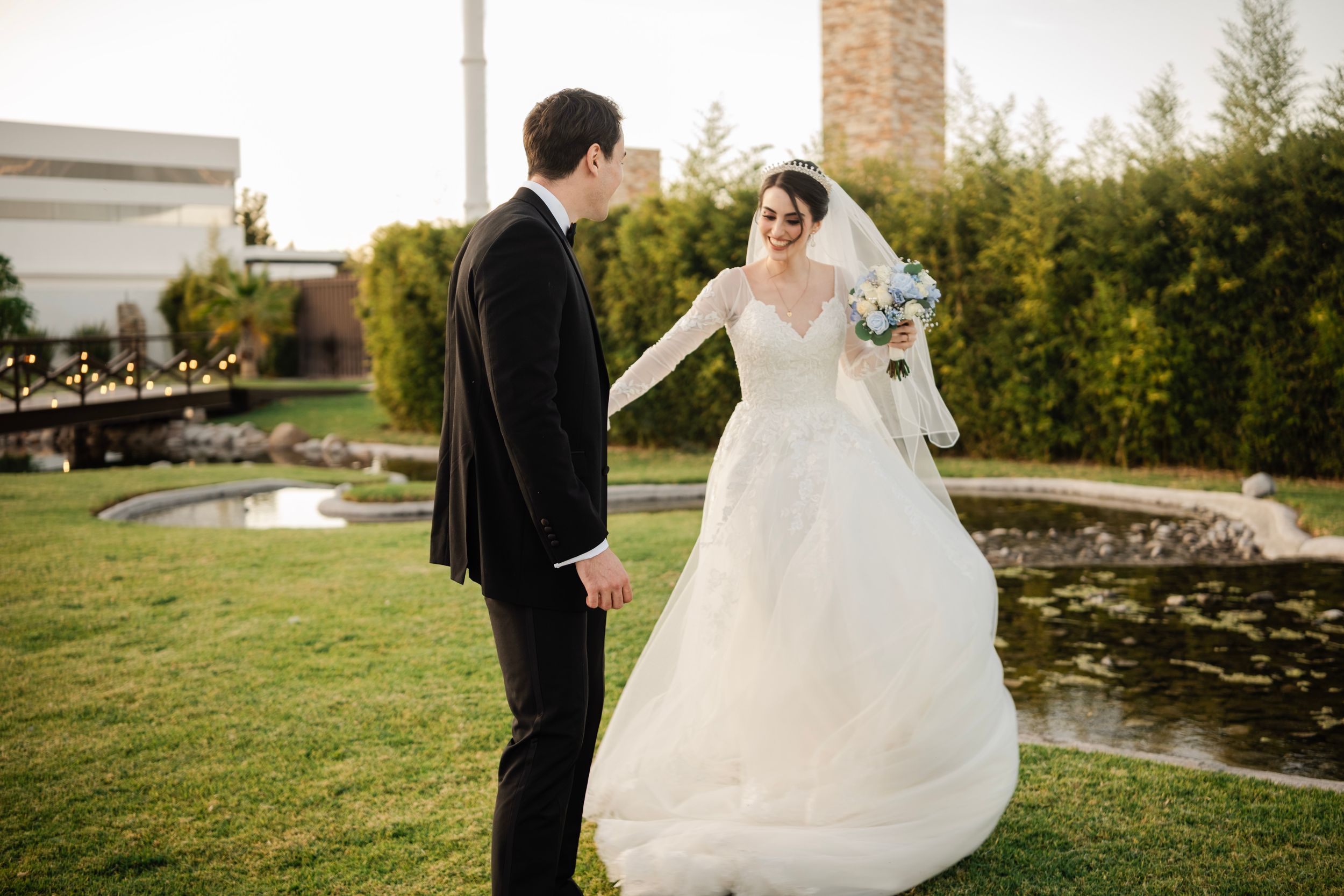 Wedding couple stands together in elegant attire by a serene pond surrounded by autumn foliage at sunset.