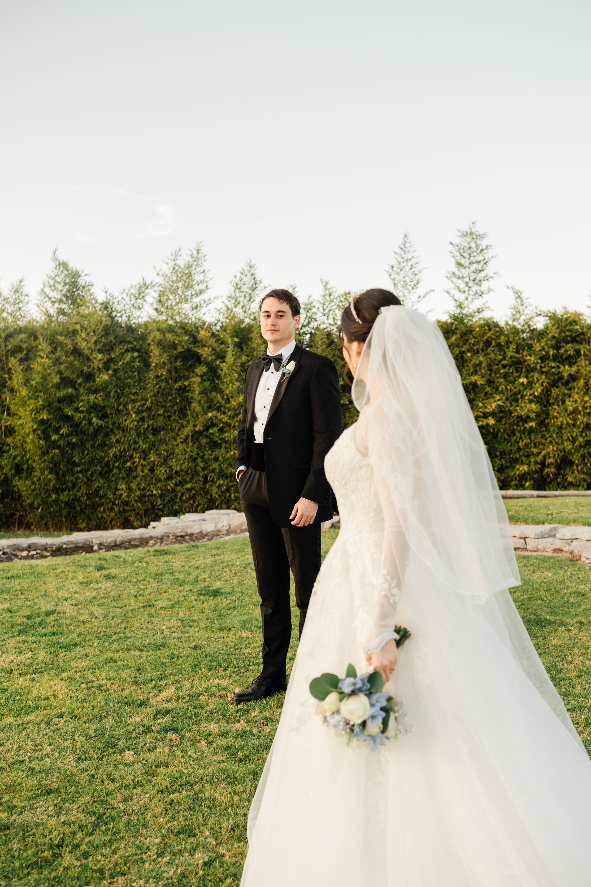A romantic wedding couple shares a tender moment on a grassy lawn with lush hedges in the background.