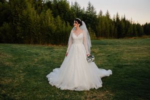 A bride in a flowing white wedding dress stands in a field with evergreen trees in the background at sunset.