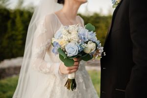 A wedding couple holds a bouquet of blue and white flowers during their outdoor ceremony.
