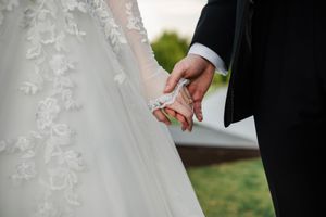 Close-up of hands holding a delicate white lace wedding dress fabric against a blurred outdoor background.