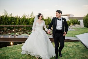 A couple in wedding attire walks together on a grassy lawn with a rustic wooden bridge in the background at sunset.
