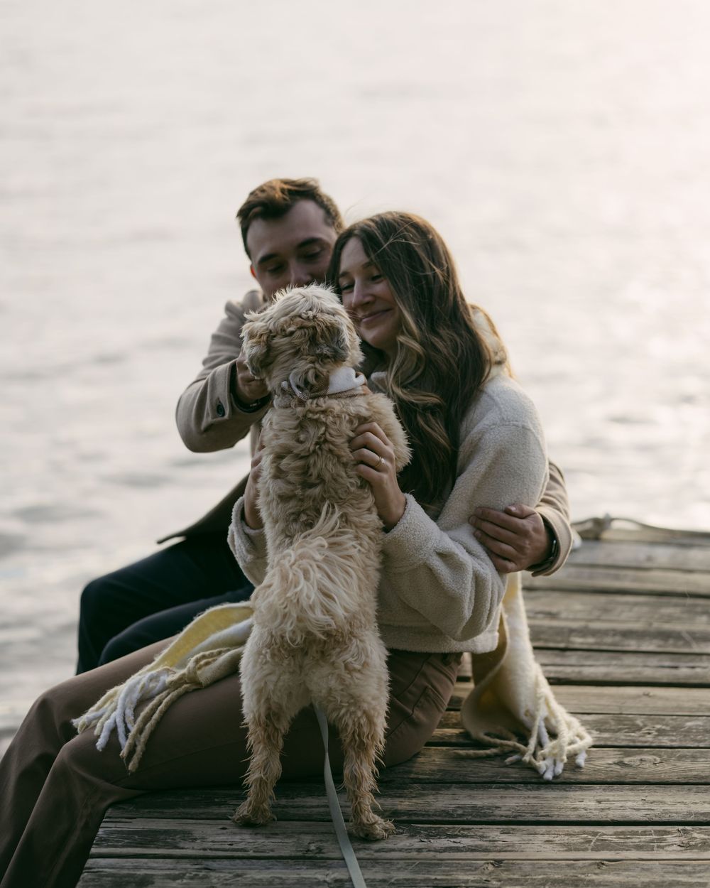 A couple sits on a wooden dock with their fluffy white dog at sunset.