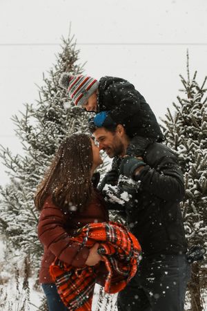 Two people in winter coats share a tender moment in the snow with evergreen trees in background.
