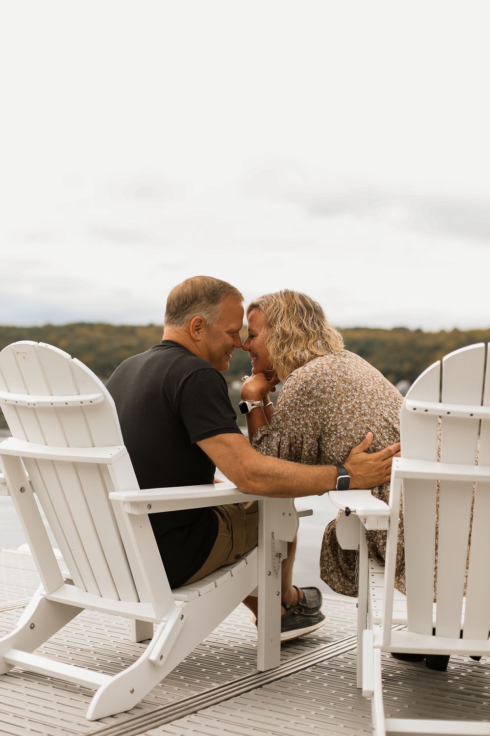 A couple shares an intimate moment while sitting in white Adirondack chairs on a wooden deck overlooking a scenic landscape.