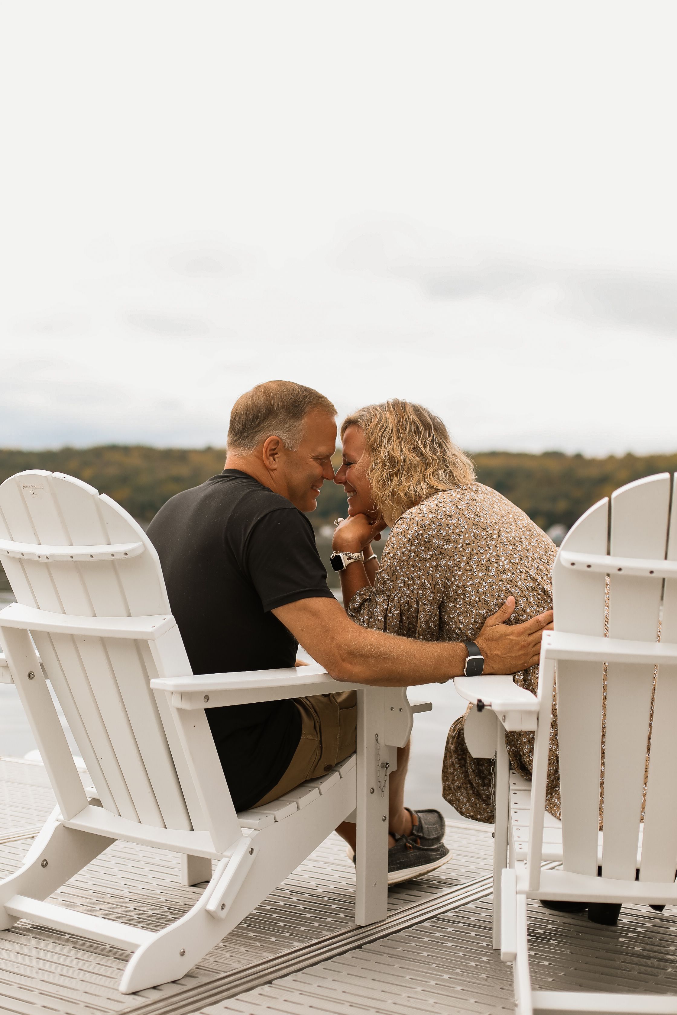 A couple shares an intimate moment while sitting in white Adirondack chairs on a wooden deck overlooking a scenic landscape.