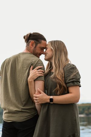 A couple embraces intimately by the water wearing casual earth-toned clothing on an overcast day.