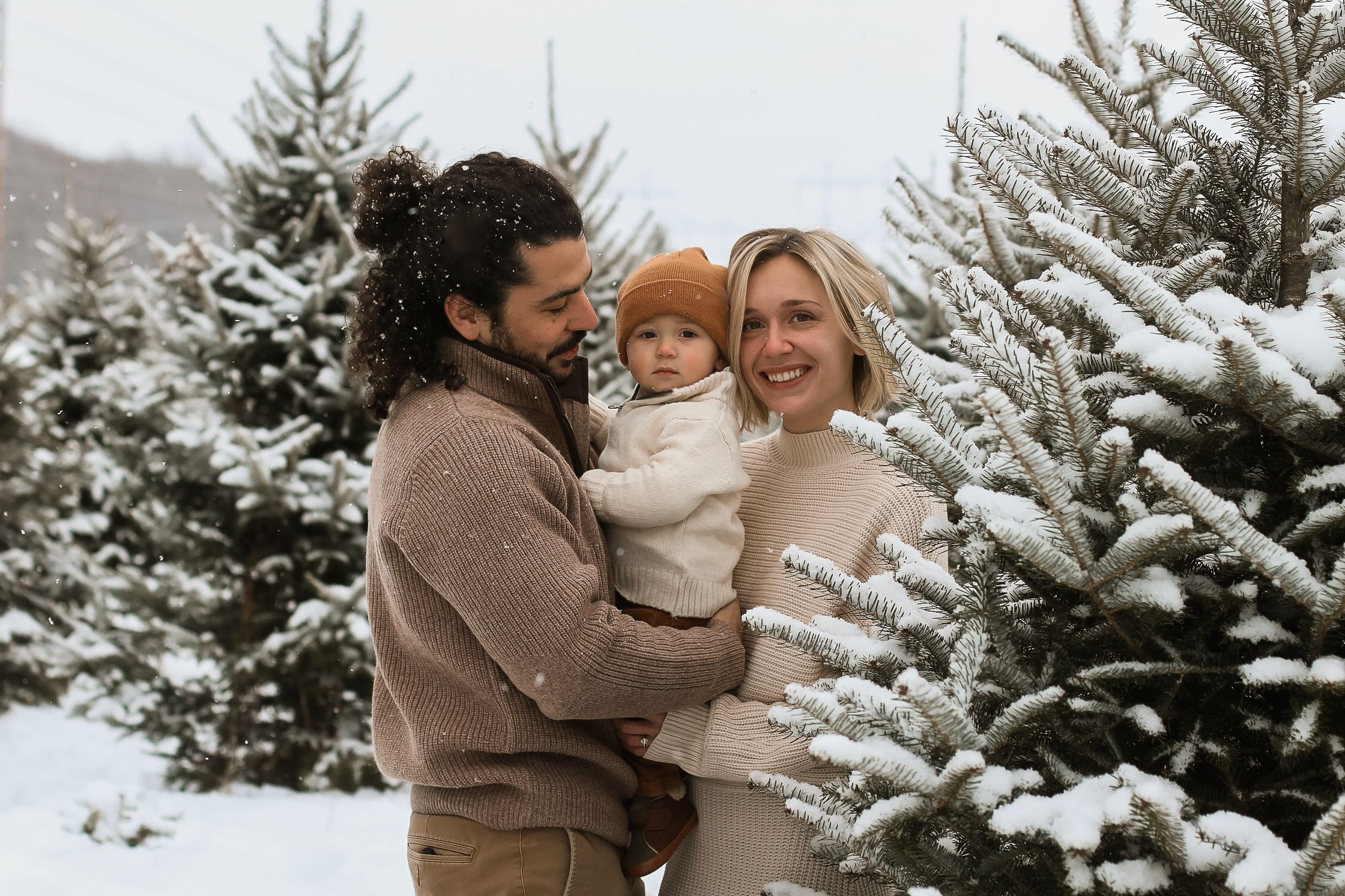A family embraces while standing among snow-covered pine trees in a wintry forest setting.