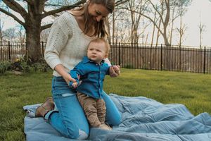 A parent and baby relax together on a blue blanket in a backyard with a wooden fence on a spring afternoon.