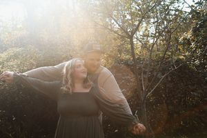 A couple shares a romantic moment in nature with golden sunlight streaming through trees behind them.