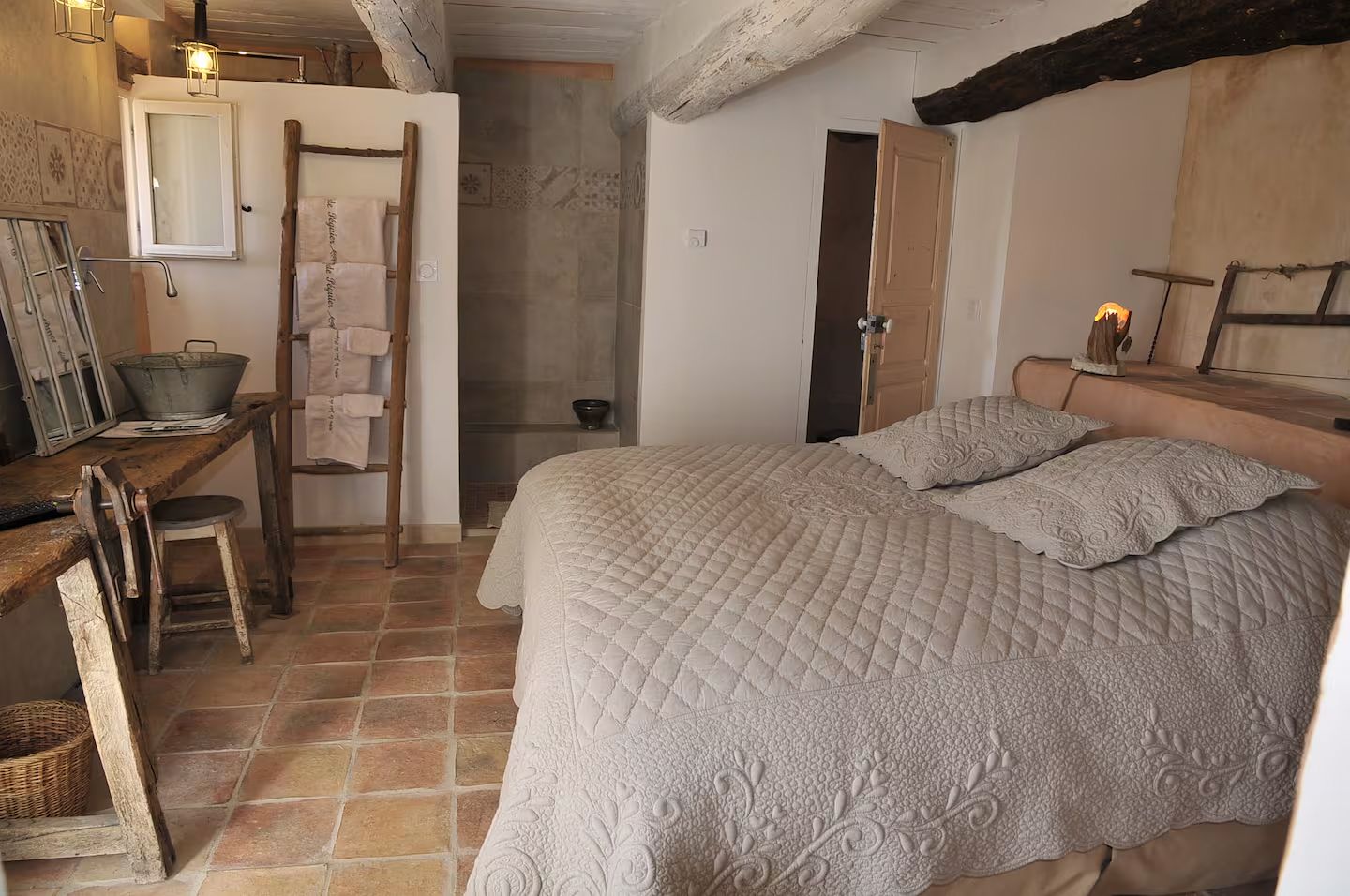Rustic bedroom interior with terracotta floor tiles, quilted white bedding, and exposed wooden beam ceiling details.