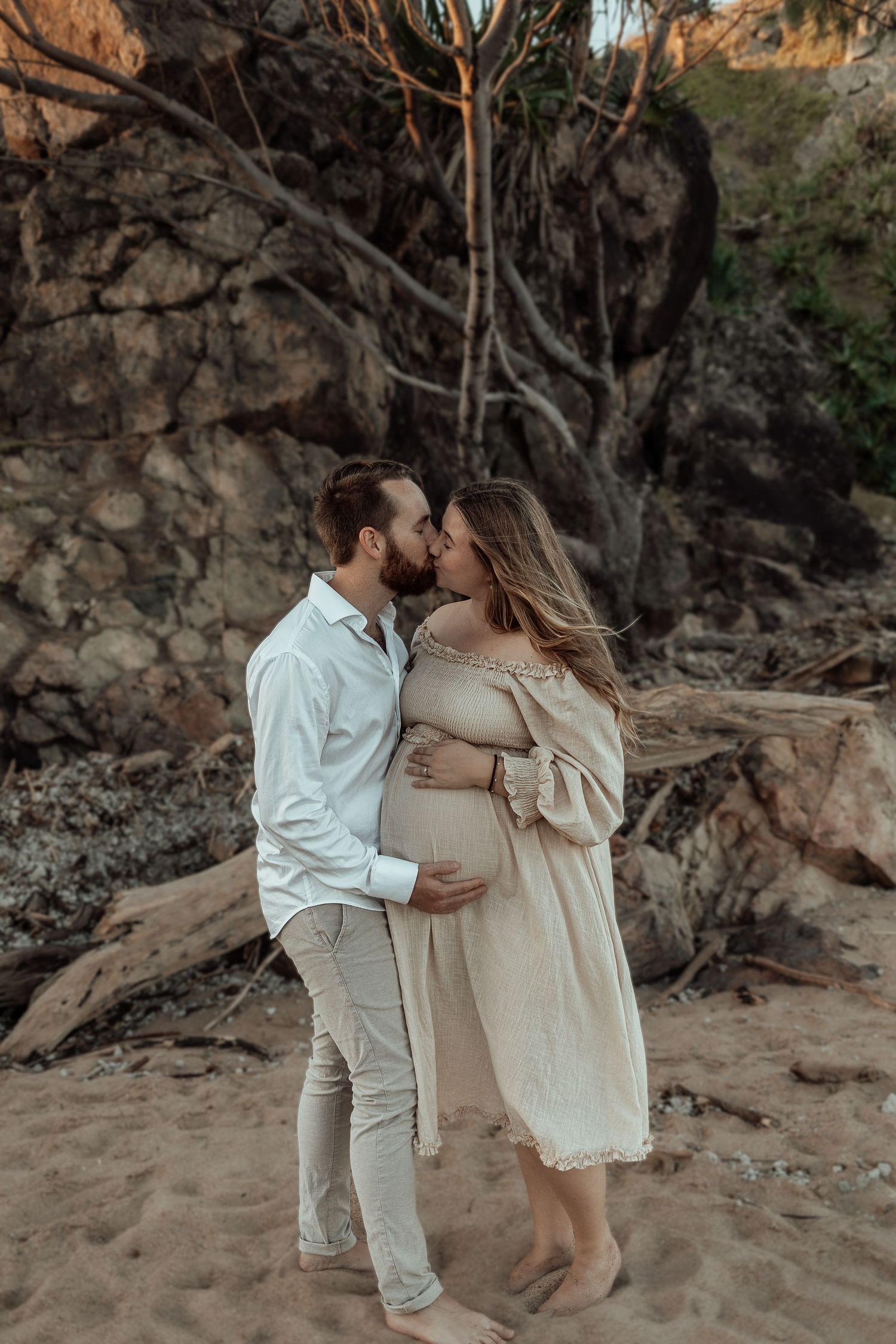 Romantic maternity photo of couple embracing on rocky beach during sunset.