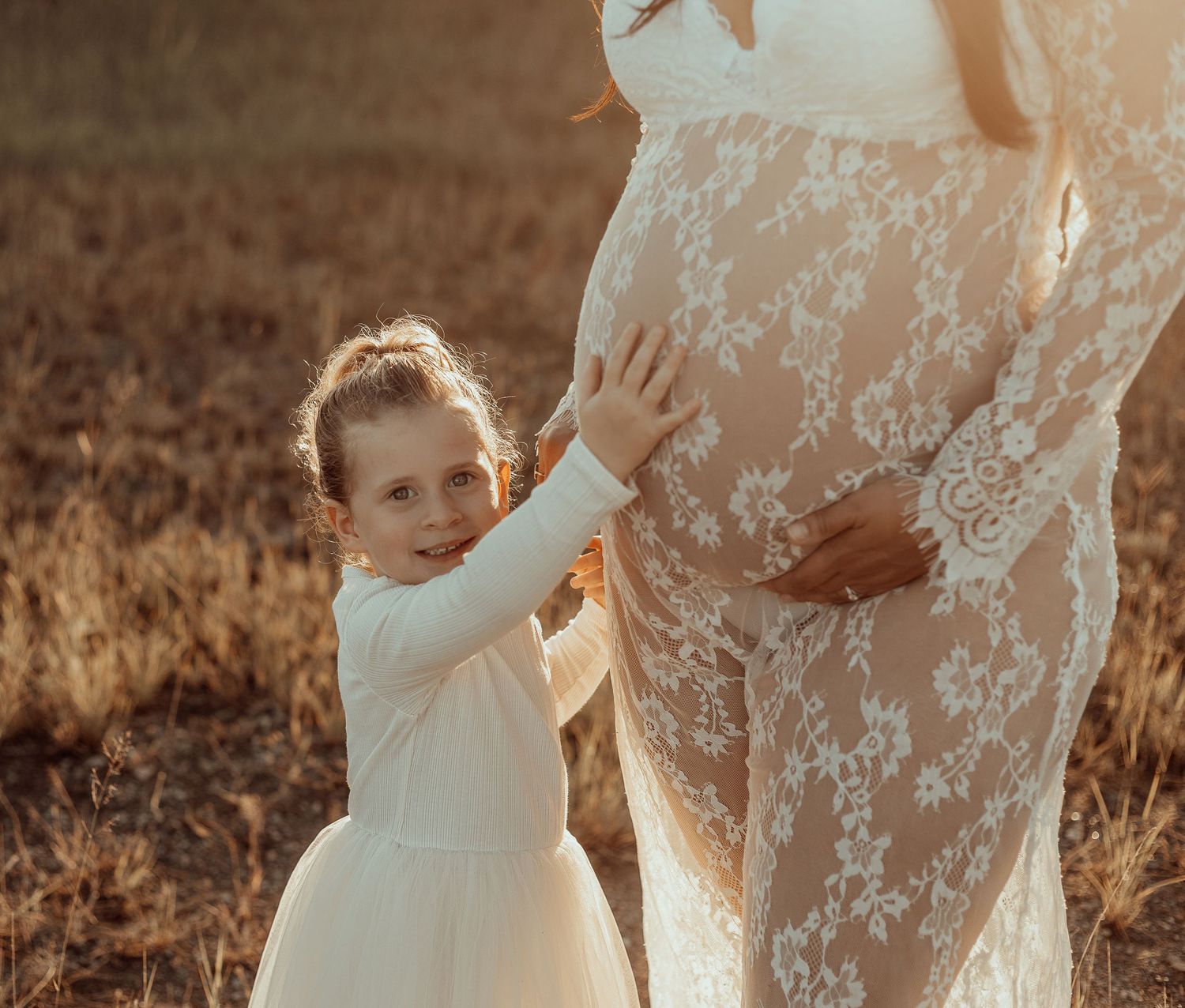 Young child in white dress reaches up to touch mother's lace maternity gown during sunset photoshoot.
