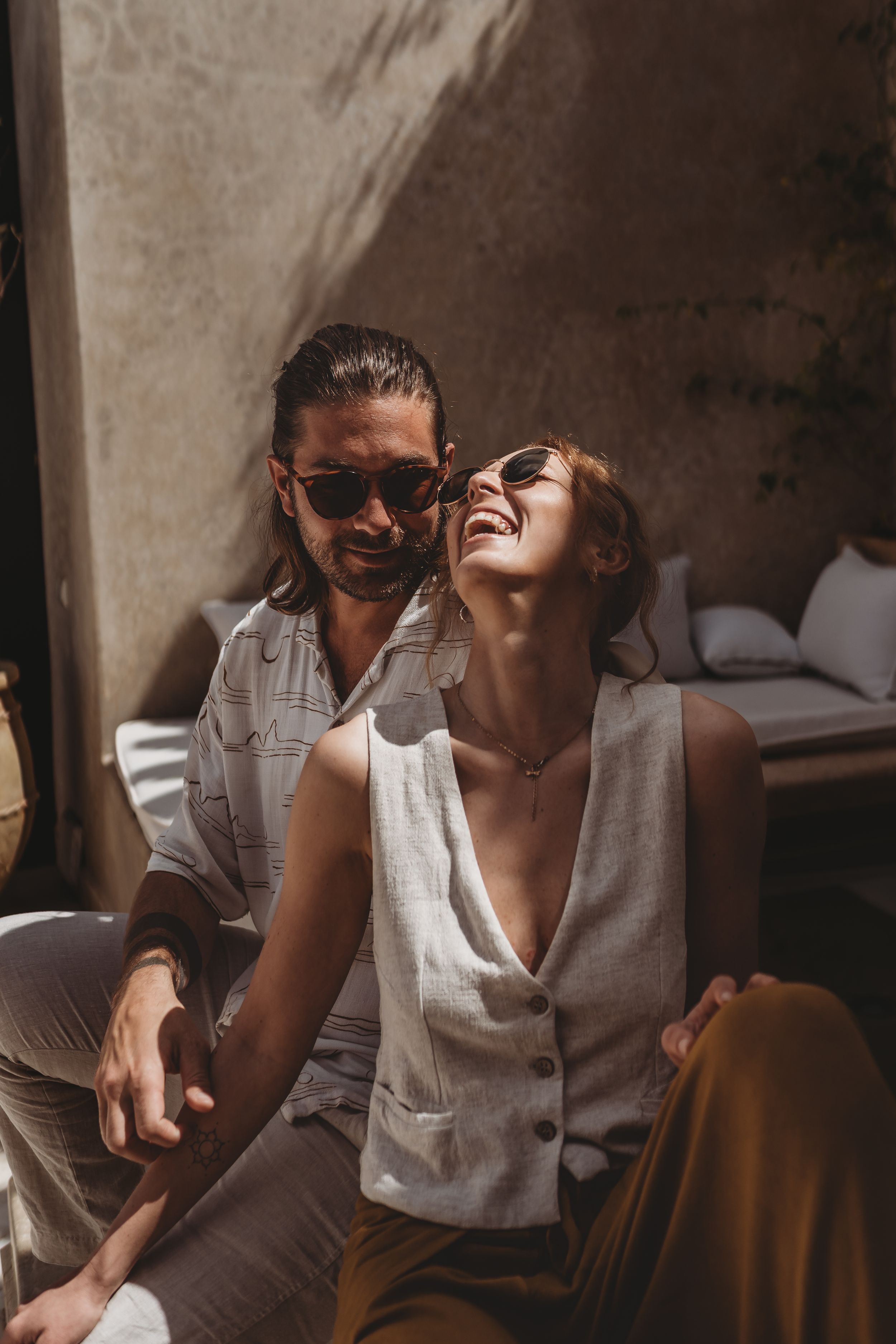 A laughing couple in casual linen outfits sit together in warm sunlight casting shadows on a wall.