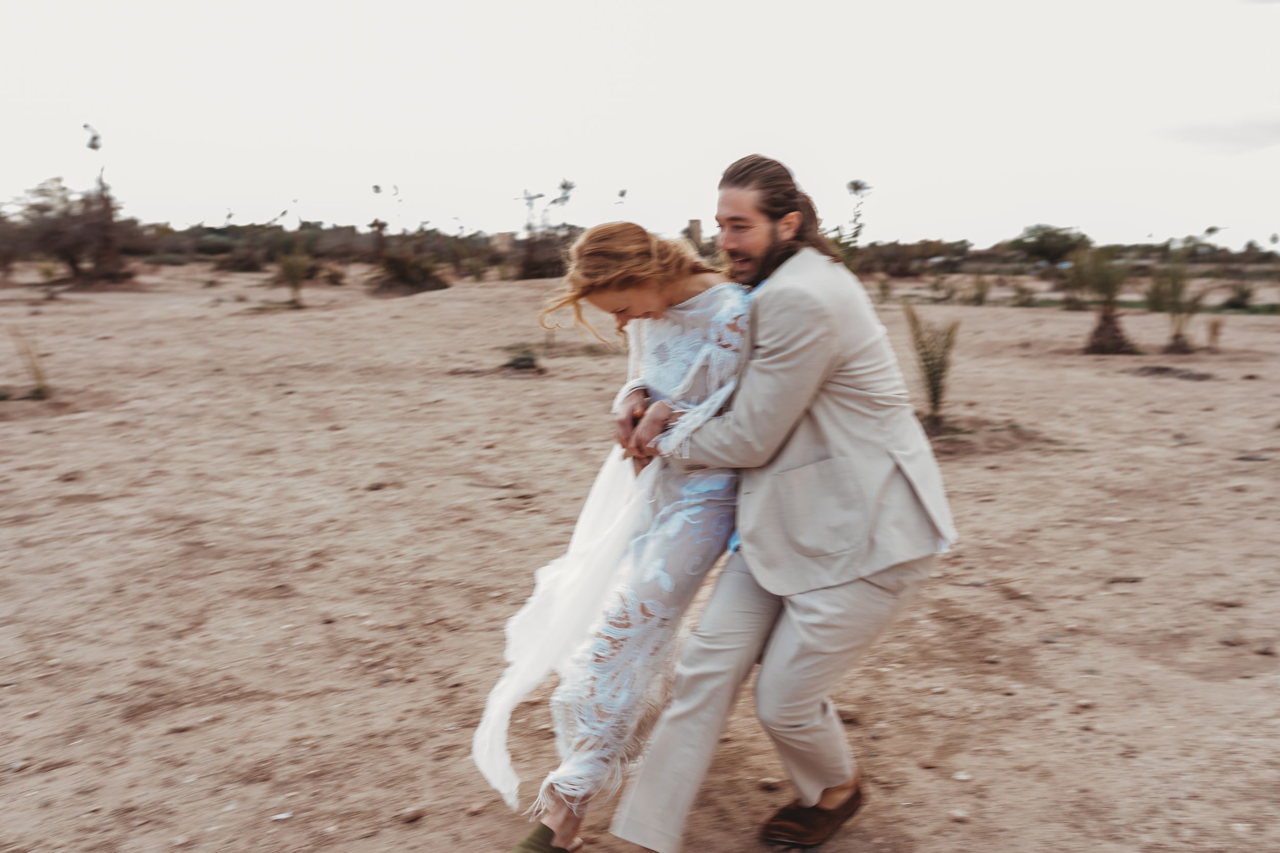 A couple in light colored outfits playfully dancing together on a sandy desert landscape at dusk.