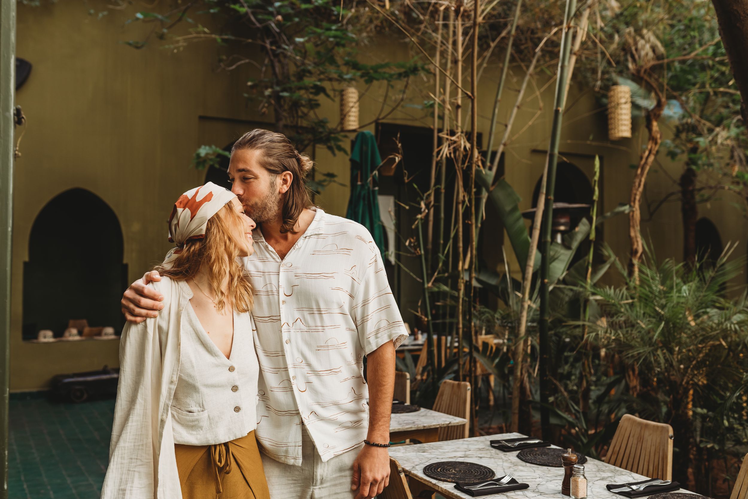 A tender embrace between two people in a lush garden patio setting with rustic furniture and natural lighting.