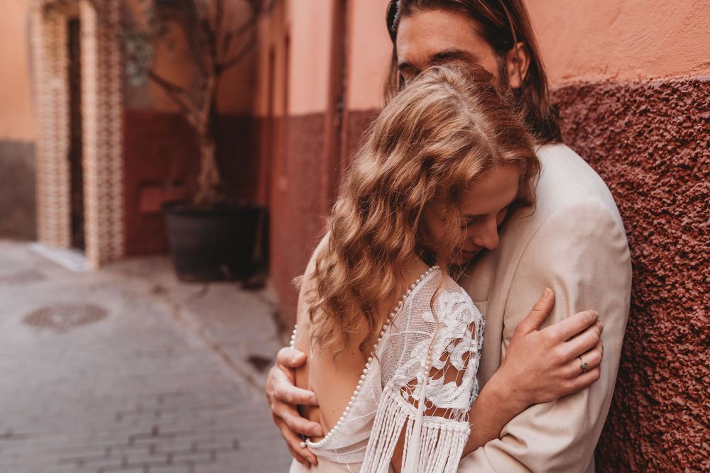Romantic couple embracing against terracotta wall in narrow cobblestone street wearing matching light colored outfits.