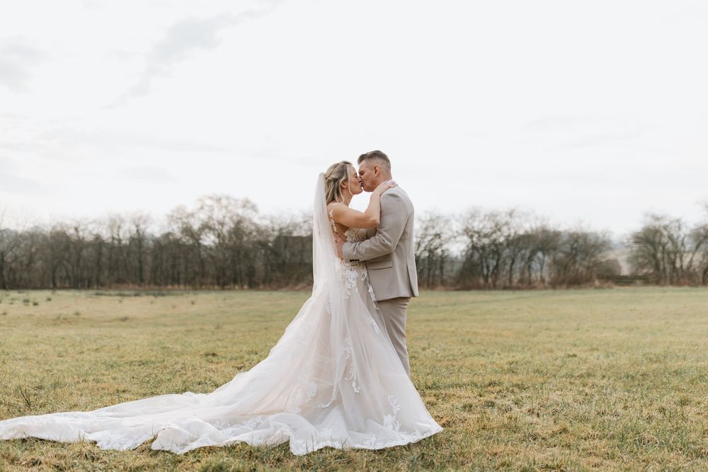 A bride in a flowing white gown and groom in gray suit embrace lovingly in a scenic countryside field at sunset.