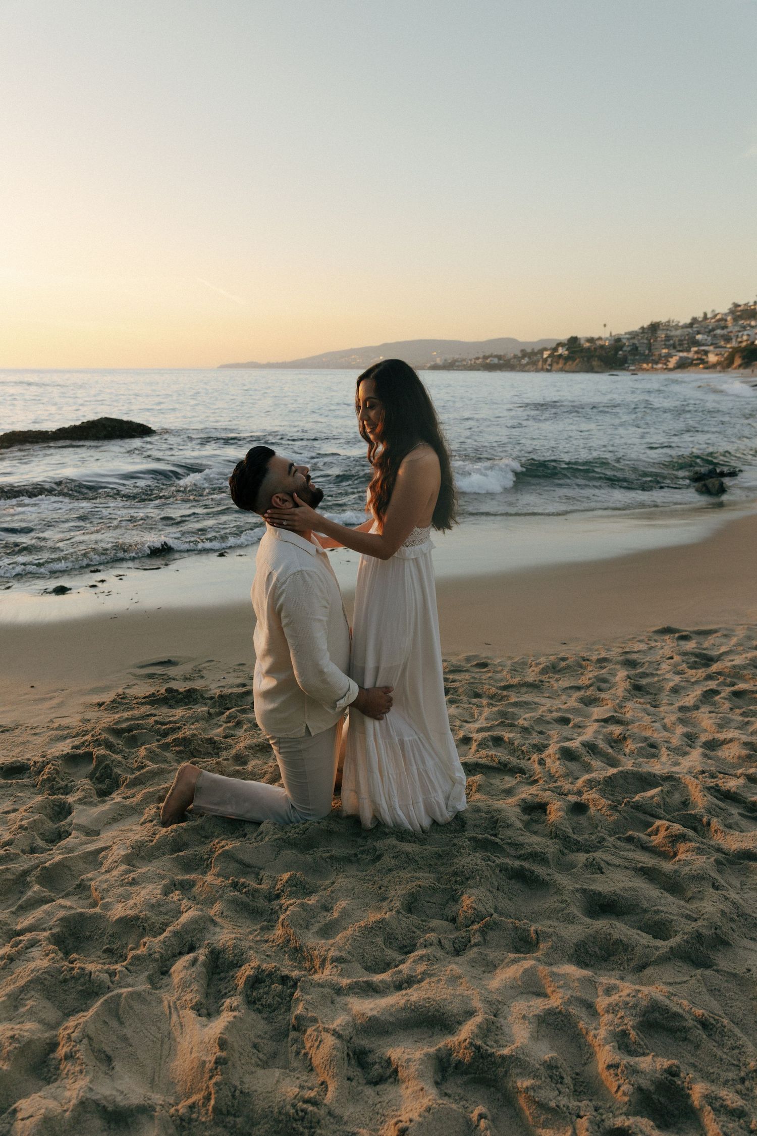 A romantic beach proposal at sunset with waves crashing on the shore with coastal mountains in the background.
