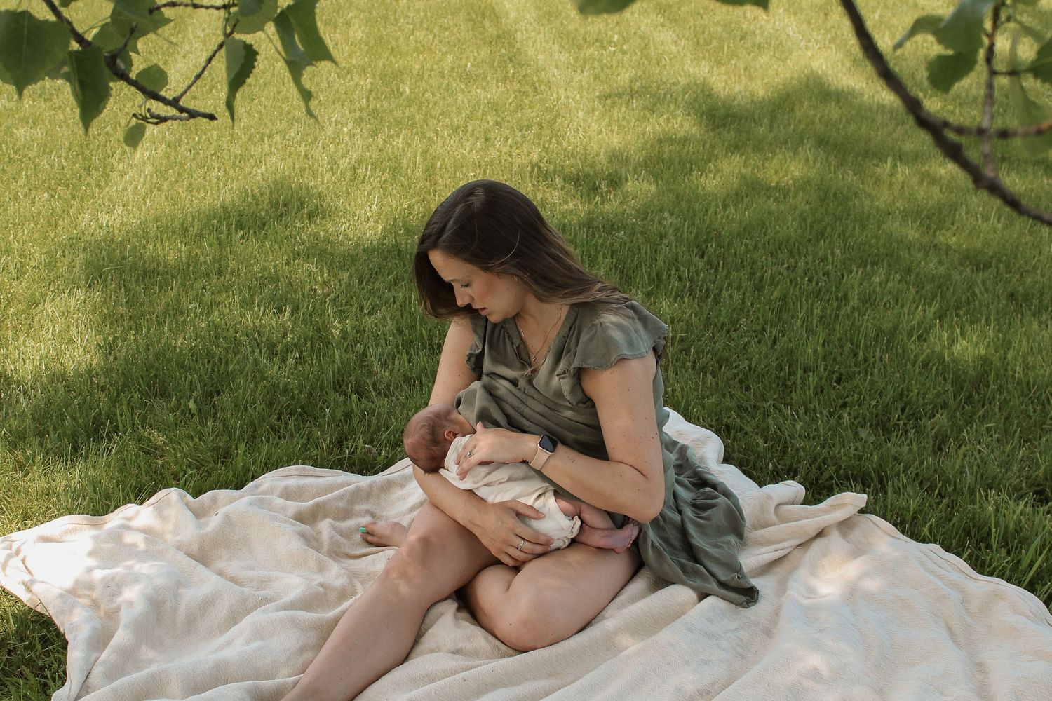 A person sits on a white blanket in dappled sunlight on grass while nursing a baby on a summer day.