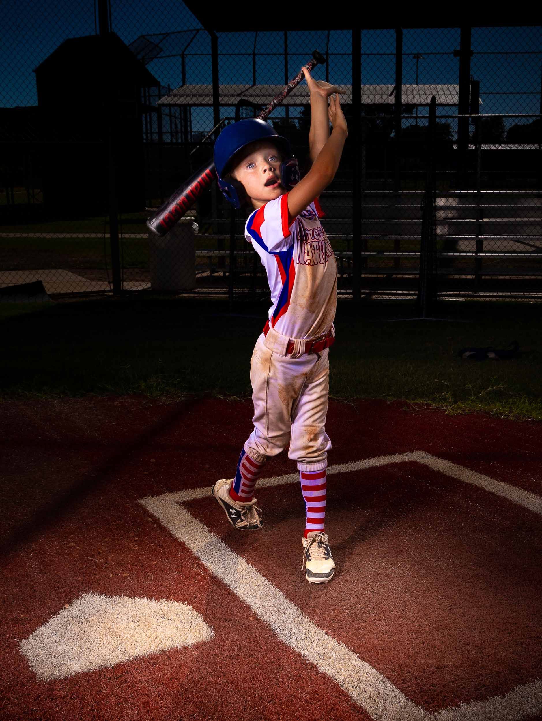 Young baseball player in patriotic uniform stands illuminated at home plate on a dusky baseball field.