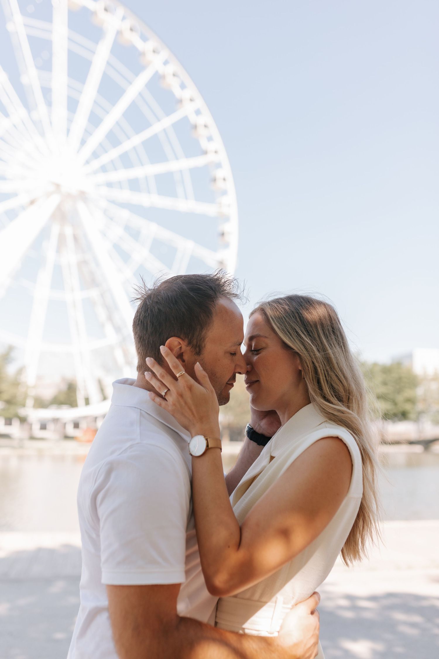 Couple sharing a tender moment about to kiss with a Ferris wheel and blue sky in the background.
