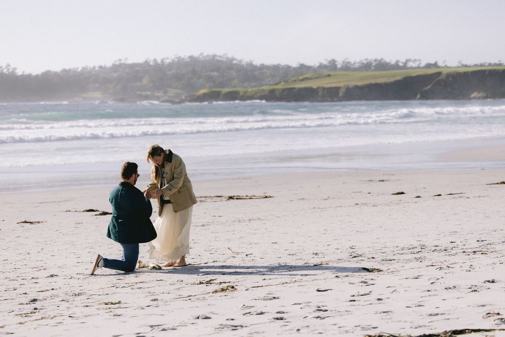 A romantic proposal scene unfolds on a serene sandy beach with crashing waves and coastal hills in the background.