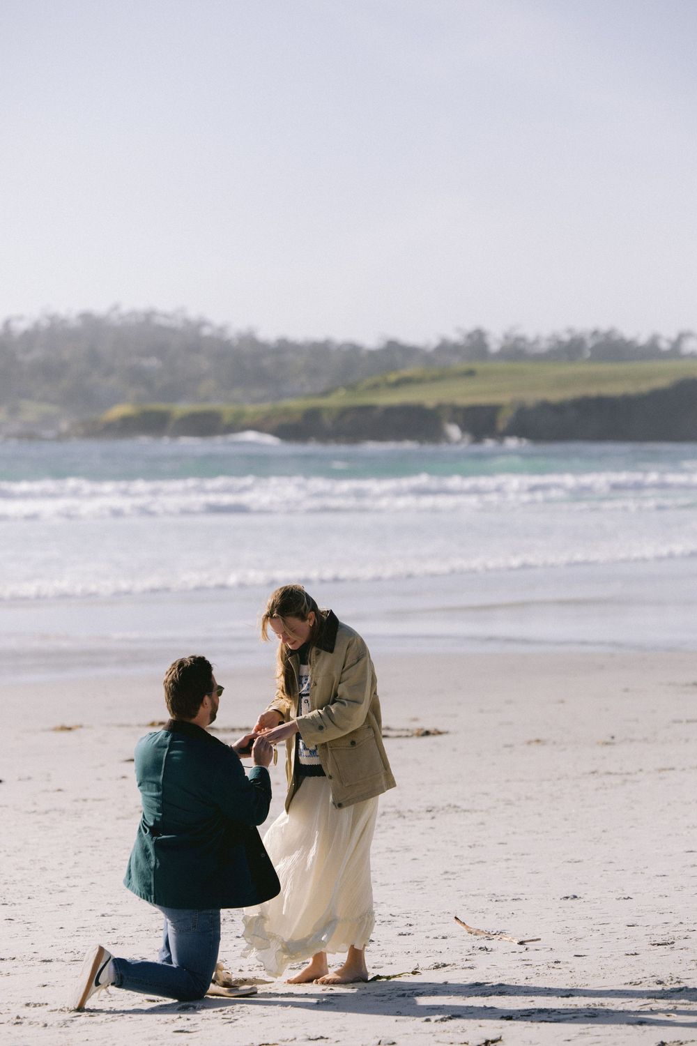 Two people share a romantic moment on a scenic beach with waves and coastal cliffs in the background.