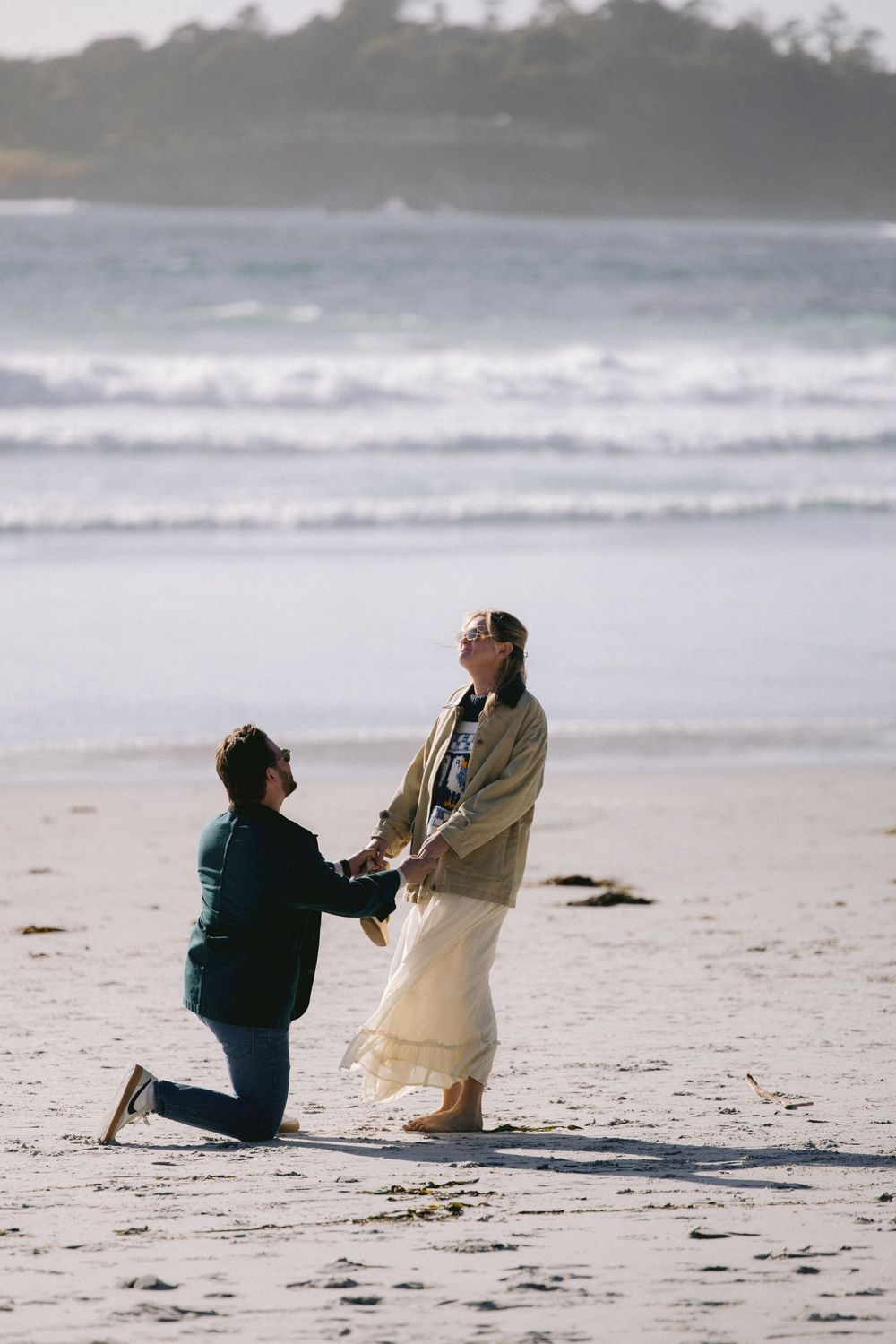 A couple shares a romantic beach proposal moment with crashing waves in the background on a misty shoreline.