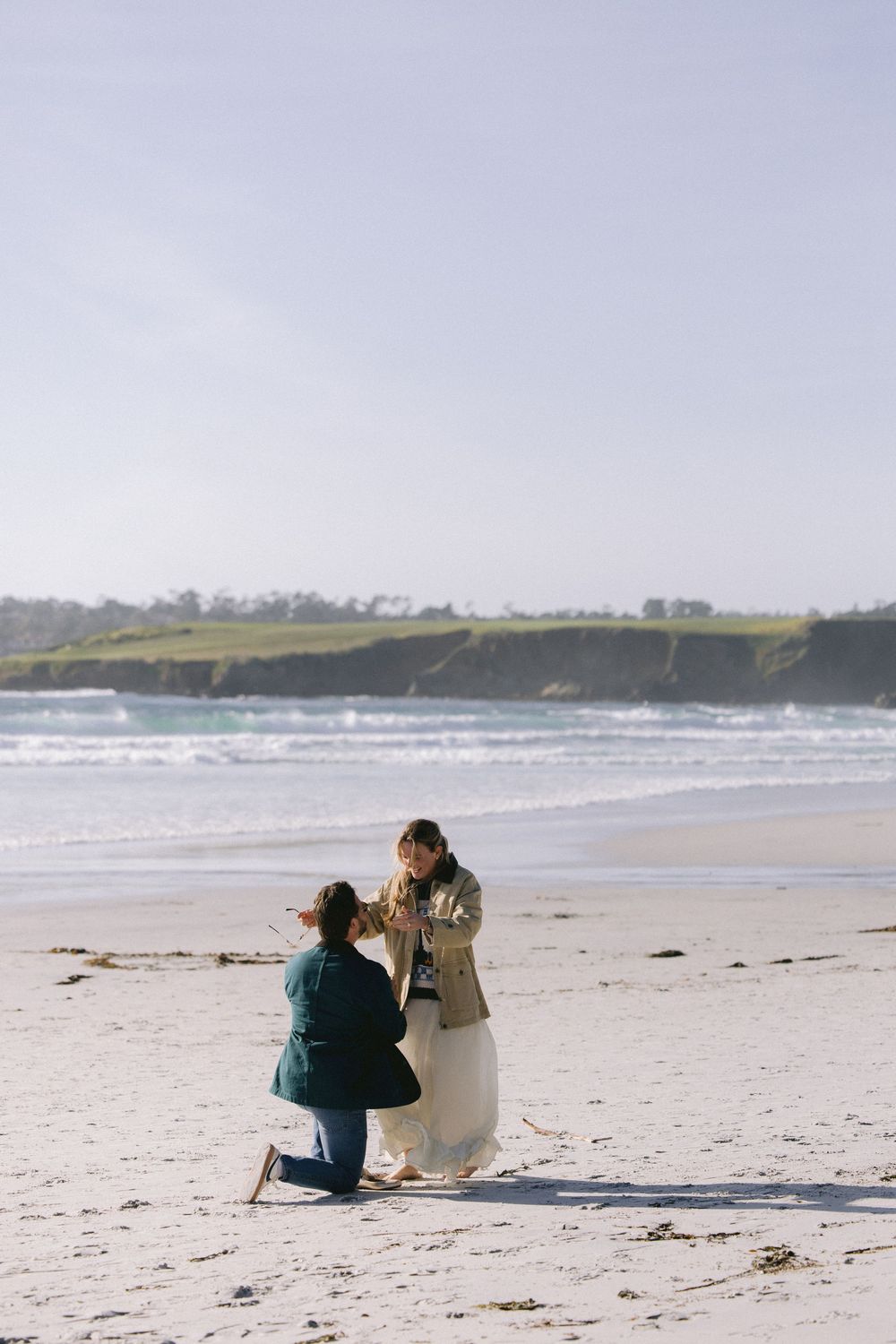 A romantic marriage proposal unfolds on a serene beach with rolling waves and green cliffs in the background.