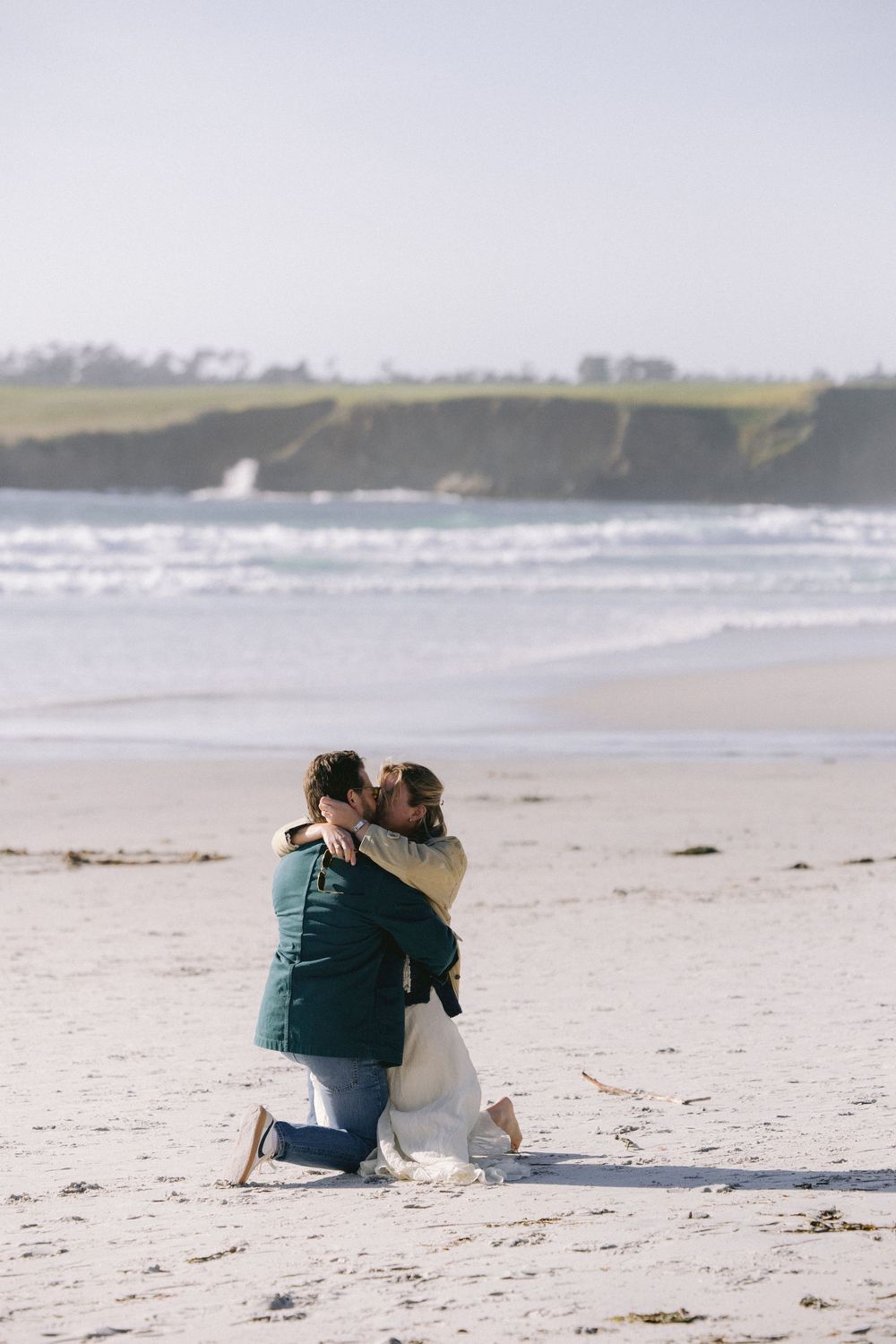 A couple embraces while kneeling on a sandy beach with rugged coastal cliffs and crashing waves in the background.