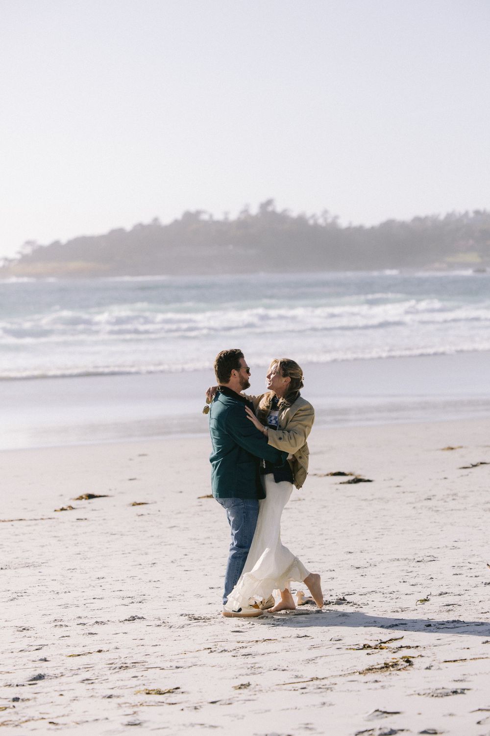 A couple embraces tenderly while standing on a misty beach with crashing waves and rocky coastline in the background.