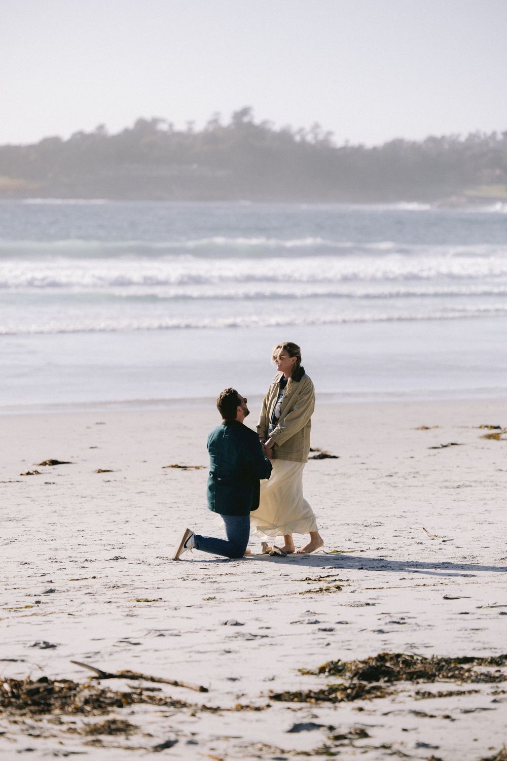 A romantic proposal scene on a misty beach with crashing waves and coastal trees in the background.