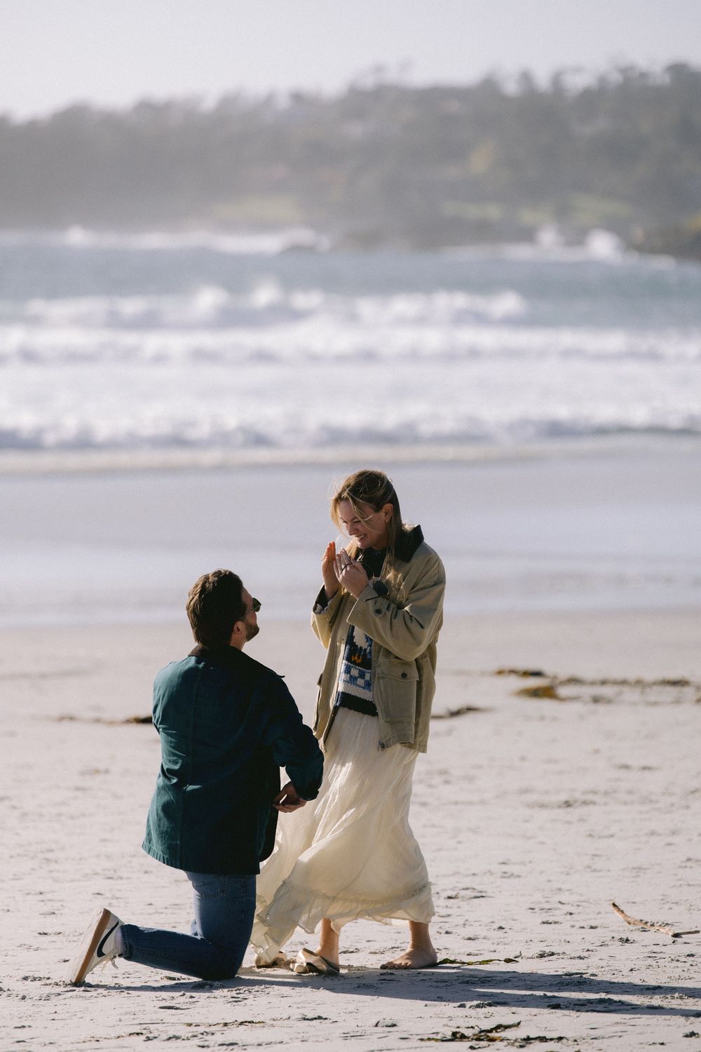 A couple shares a romantic proposal moment on a misty beach with crashing waves in the background.