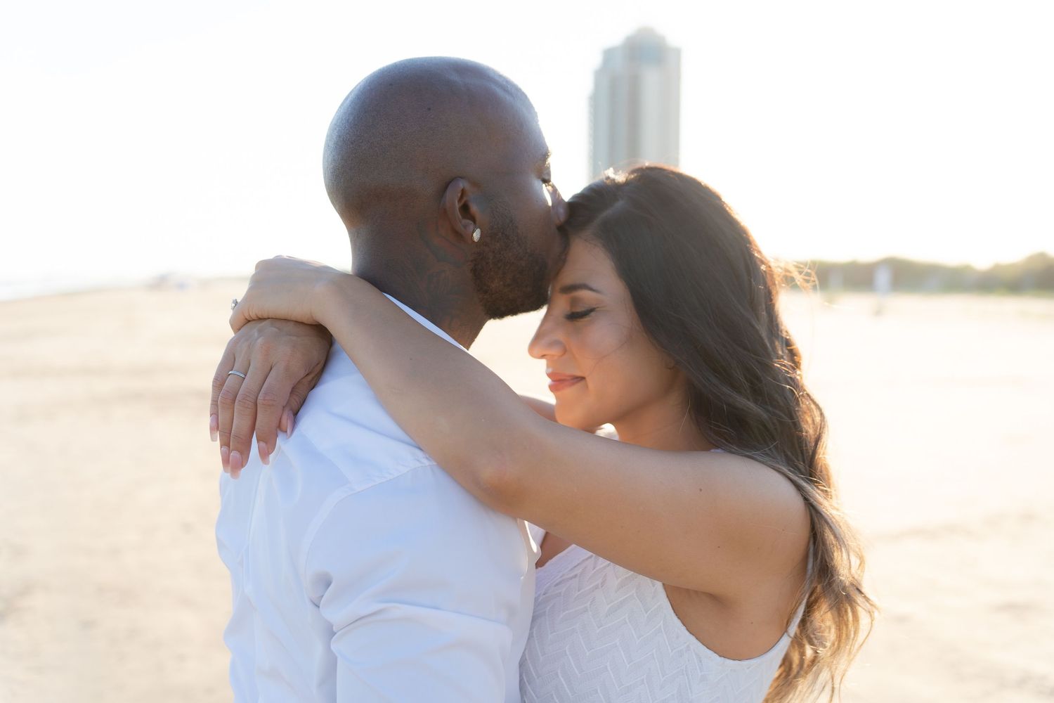 A romantic couple embraces on a beach at sunset, the bride in white and her partner in a white shirt.