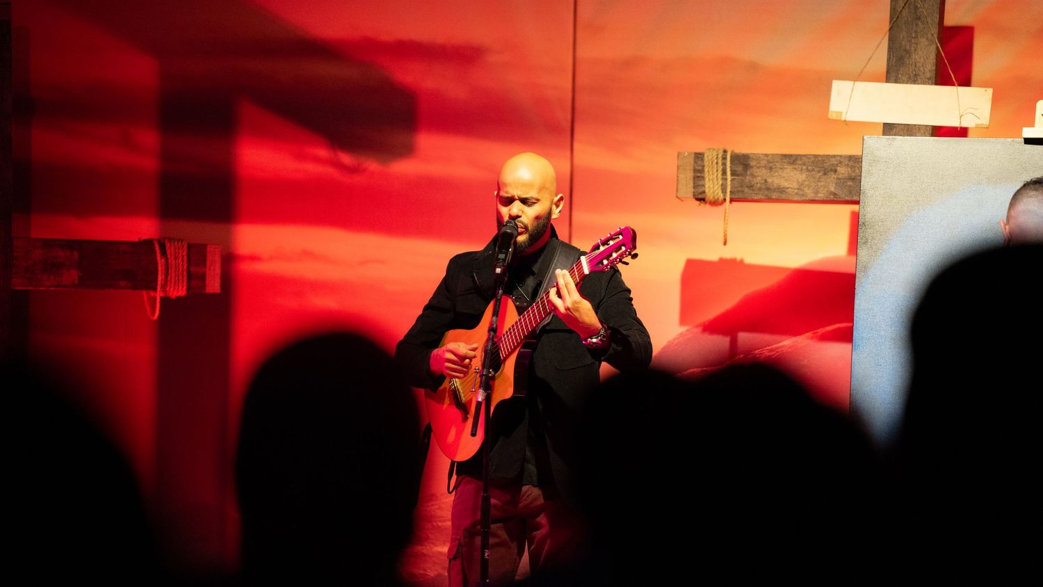 A performer plays acoustic guitar on stage against a dramatic red sunset backdrop during a live concert performance.