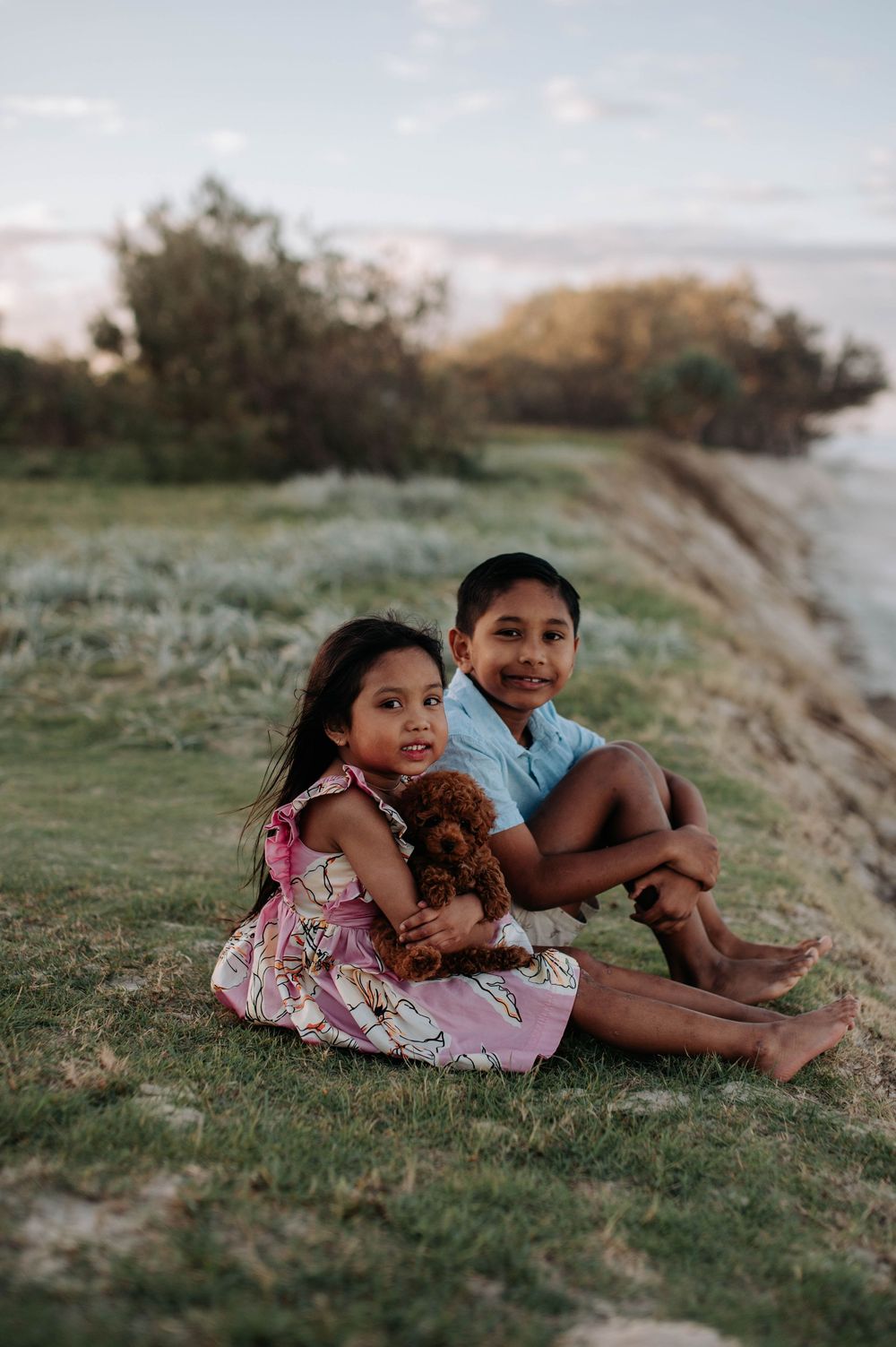 Two young children sitting on grassy hill at sunset with a small brown puppy in a peaceful outdoor setting.