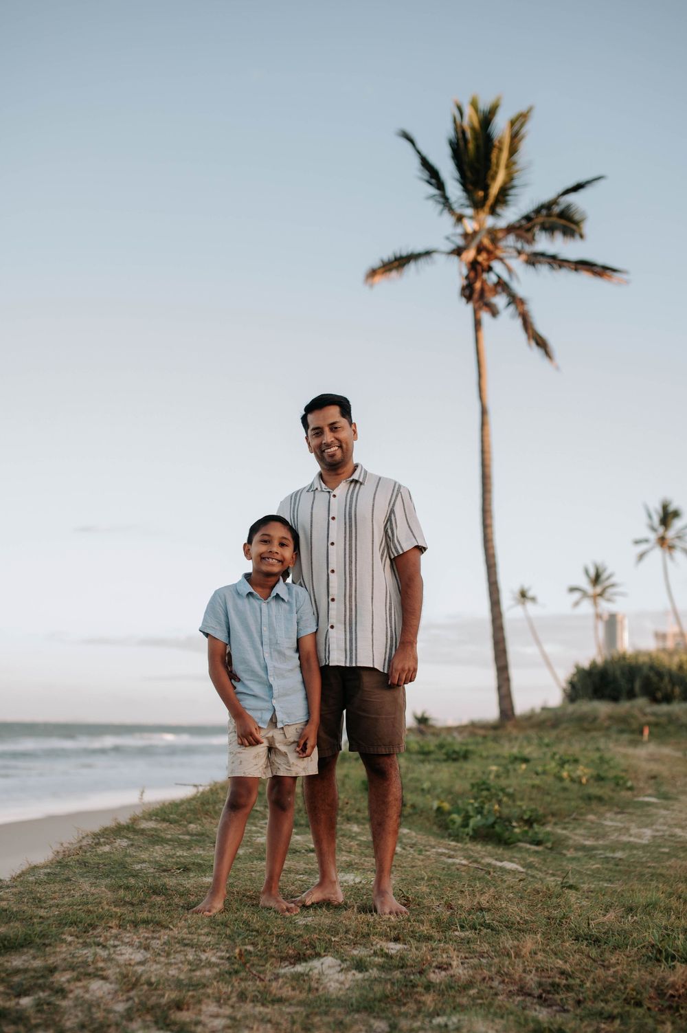 Two people stand barefoot on a grassy oceanfront hill with a tall palm tree against a sunset sky in Hawaii.